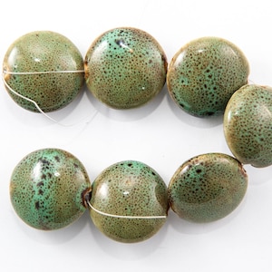 Könnte beinhalten: Sieben runde, gesprenkelte Perlen in Grün- und Brauntönen sind mit weißem Faden aufgefädelt. Die Perlen haben eine glänzende Oberfläche und eine leicht abgeflachte Form. Die Perlen sind wahrscheinlich für die Schmuckherstellung.