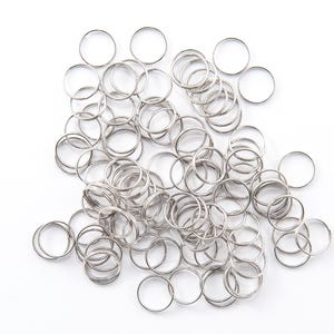 May include: A collection of small, silver-colored metal rings scattered on a white surface. The rings are circular and appear to be made of a shiny, metallic material. The image is a close-up shot, highlighting the details of the rings.