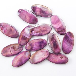 May include: A collection of oval-shaped beads in shades of purple and pink, scattered on a white surface. The beads have a glossy, iridescent finish, with visible natural patterns and textures. Each bead has a small hole for stringing.