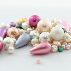 Może przedstawiać: Kolekcja różnorodnych koralików w różnych kształtach i rozmiarach. Koraliki są w odcieniach różu, bieli, fioletu i opalizujących kolorów. Niektóre są okrągłe, inne w kształcie łezki.