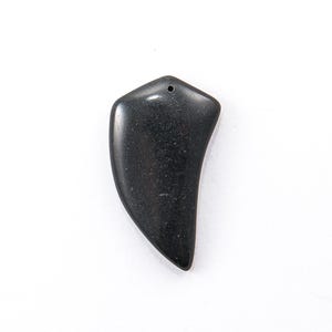Puede incluir: Un colgante negro pulido en forma de garra. El colgante tiene una superficie lisa y brillante y un pequeño agujero en la parte superior para una cadena. El diseño es curvo y cónico, con una parte superior redondeada y una inferior puntiaguda.