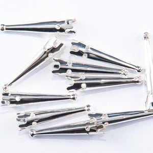 May include: A collection of silver-colored metal hair clips, arranged on a white surface. Each clip has a tapered design with a rounded end and a toothed section for gripping hair. The clips are approximately 2 inches in length.