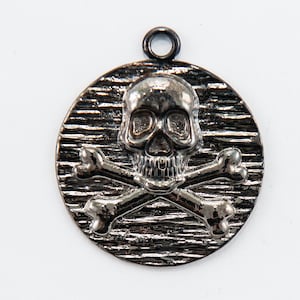 May include: A silver-toned pendant featuring a skull and crossbones design. The skull has a detailed face with eye sockets, a nose, and a mouth. The crossbones are positioned behind the skull, creating a classic pirate symbol.