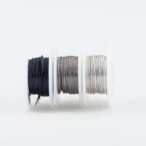 May include: Three spools of thread, one black, one gray, and one white, are wound on white plastic spools.