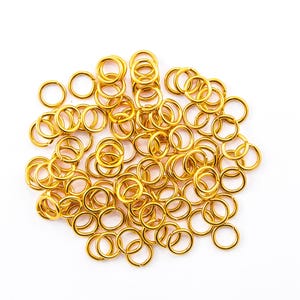 May include: A pile of small, gold-coloured jump rings on a white background. These open rings are circular and appear to be made of metal. They are commonly used in jewellery making and other crafts.