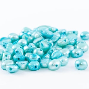 May include: A pile of light blue, irregularly shaped beads with a pearlescent finish. The beads vary in size and shape, with some having a more rounded form and others being flatter. The beads are scattered on a white background.