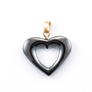 Puede incluir: Un colgante en forma de corazón con una fianza dorada. El colgante es de color gris oscuro metálico y tiene un centro hueco, creando un corazón dentro de un corazón. El colgante probablemente está hecho de metal pulido.