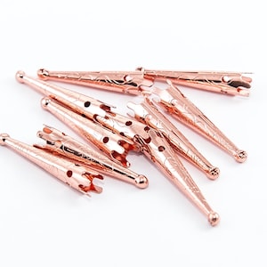 May include: A collection of rose gold hair clips, each with a decorative floral pattern and a rounded end. The clips are arranged on a white surface, showcasing their intricate design and metallic sheen.