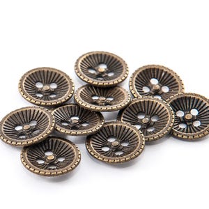 May include: A collection of bronze-colored, round buttons with a detailed, radial design. Each button has three holes for sewing. The buttons are scattered on a white background, showcasing their intricate texture and vintage aesthetic.