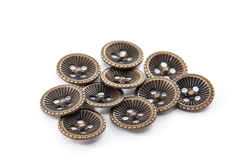 May include: A collection of bronze-colored, round buttons with a detailed, radial design. Each button has three holes for sewing. The buttons are scattered on a white background, showcasing their intricate texture and vintage aesthetic.