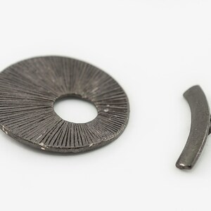 Puede incluir: Dos piezas de joyería de metal, una es un disco redondo con un agujero en el centro y una superficie texturizada, la otra es una pieza curva con una superficie lisa.