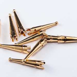 May include: Several gold-coloured metal findings, possibly jewellery components, are arranged on a white surface. The findings have a conical shape with a decorative, open-work end and a rounded tip. They appear to be new and unused.