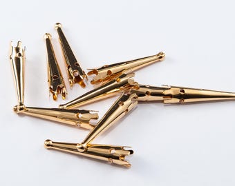 May include: A collection of gold-colored metal findings, likely jewelry components, scattered on a white surface. The findings have a conical shape with a decorative, open-work end and a rounded tip. They appear to be new and unused.