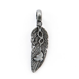 Pode incluir: Um pingente de metal prateado em forma de pena com um pequeno desenho de pássaro. O pingente tem um laço na parte superior para prender a uma corrente.