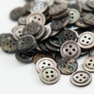 May include: A pile of round, dark gray buttons with four holes each. The buttons have a pearlescent sheen and varying shades of gray and brown. They are scattered on a white surface, creating a textured appearance. These buttons are ideal for sewing and crafts.