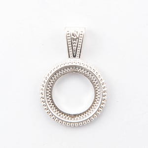 May include: A silver pendant with a circular design, featuring a textured edge and a decorative bail. The pendant is set against a white background, highlighting its intricate details and craftsmanship. The pendant is likely designed to hold a coin or other small item.