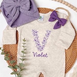 May include: A beige baby onesie with a purple floral letter V and the name "Violet" embroidered on the front. The onesie is paired with a purple turban hat and a purple bow headband.