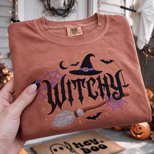 May include: A terracotta-colored crewneck sweatshirt featuring black embroidered text that reads "WITCHY" in a stylized font. The design includes a black witch hat with a purple band, a broomstick, purple spiderwebs, bats, and stars.