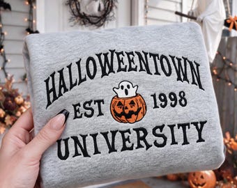 Może przedstawiać: Jasnoszara bluza z okrągłym dekoltem z czarnym haftem o treści HALLOWEENTOWN UNIVERSITY oraz EST 1998. Pośrodku między napisami znajduje się wyhaftowana grafika przedstawiająca małego białego duszka siedzącego wewnątrz pomarańczowej dyni.