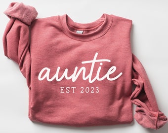 May include: A pink sweatshirt with the text "auntie est 2023" in white.