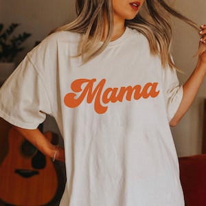 May include: A white oversized t-shirt with the word "Mama" printed in a retro orange font.