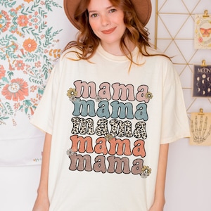 May include: A white t-shirt with the text "mama mama mama mama" in a retro font with different colors and patterns. The text is stacked vertically with a floral design on either side.