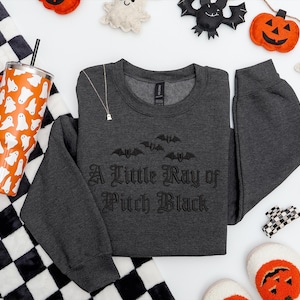May include: A dark gray sweatshirt with the text "A Little Ray of Pitch Black" and bat graphics. An orange tumbler with white ghost designs, a black and white checkered blanket, and Halloween decorations are also visible.