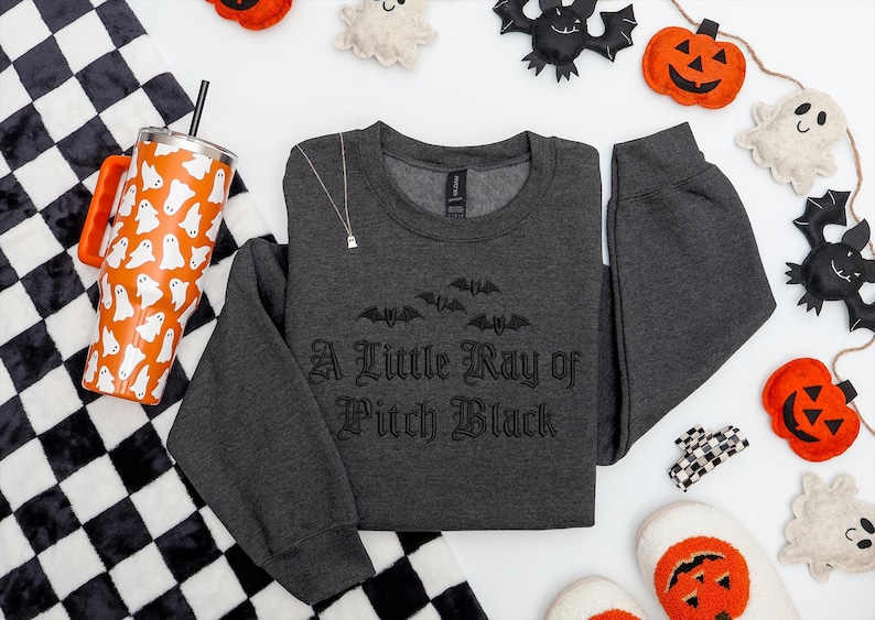 May include: A dark gray sweatshirt with the text "A Little Ray of Pitch Black" and bat graphics. An orange tumbler with white ghost designs, a black and white checkered blanket, and Halloween decorations are also visible.
