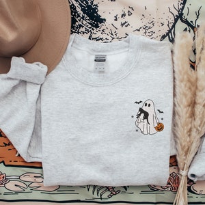 Op de afbeelding: Een grijs sweatshirt met een zwart-witte illustratie van een geest die een zwarte kat en een pompoen vasthoudt. De geest wordt omringd door vleermuizen.