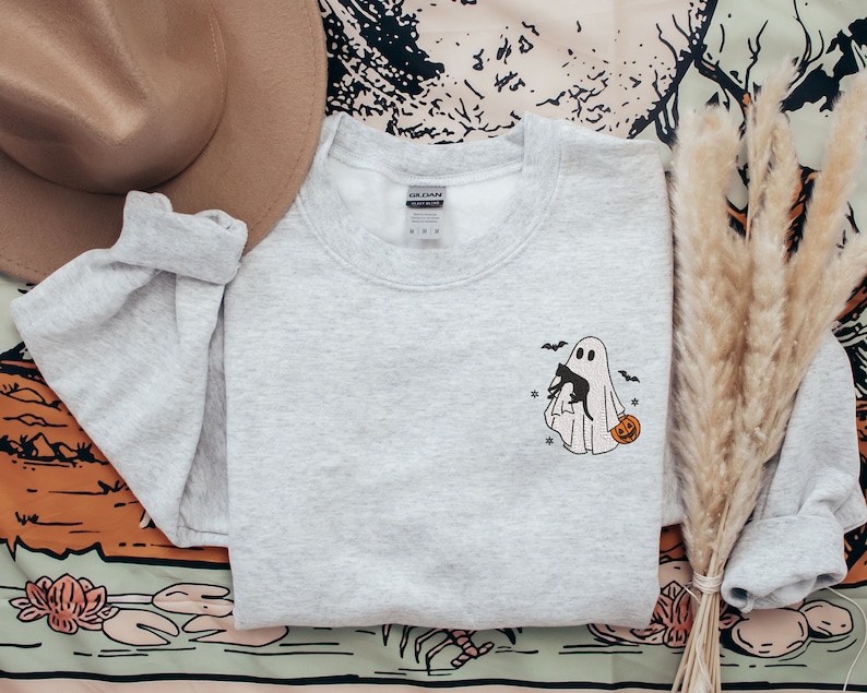Op de afbeelding: Een grijs sweatshirt met een zwart-witte illustratie van een geest die een zwarte kat en een pompoen vasthoudt. De geest wordt omringd door vleermuizen.