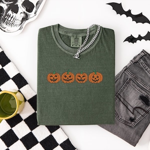 Pode incluir: Uma t-shirt verde-oliva com um gráfico bordado de quatro abóboras cor de laranja com expressões faciais distintas, dispostas numa fila horizontal ao centro. A t-shirt tem um decote redondo e uma etiqueta da marca Comfort Colors.