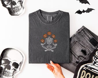 May include: A charcoal gray t-shirt featuring an embroidered white skeleton sitting in a cross-legged meditation pose, juggling four orange jack-o'-lantern pumpkins. The shirt has a crew neckline and a Comfort Colors brand label at the collar.