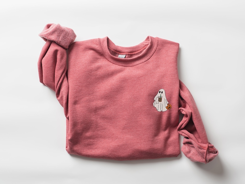 May include: A pink sweatshirt with a white ghost holding a cup of coffee and a small pumpkin. The ghost is embroidered on the front of the sweatshirt.