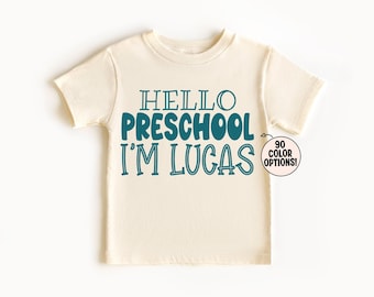 Puede incluir: Una camiseta de manga corta de color crema con texto en azul verdoso oscuro que dice HELLO PRESCHOOL I'M LUCAS. Un pequeño gráfico circular a la derecha contiene una flecha que apunta al texto con las palabras 90 COLOR OPTIONS dentro.