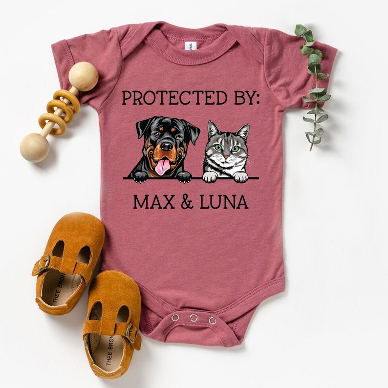 以下が含まれることがあります： 「PROTECTED BY: MAX & LUNA」という文字と、ロットワイラーと猫の漫画が描かれた、マルーン色のベビーロンパースです。