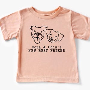 A light pink t-shirt with a graphic design featuring two dog faces and the text "Sora & Odin's NEW BEST FRIEND".