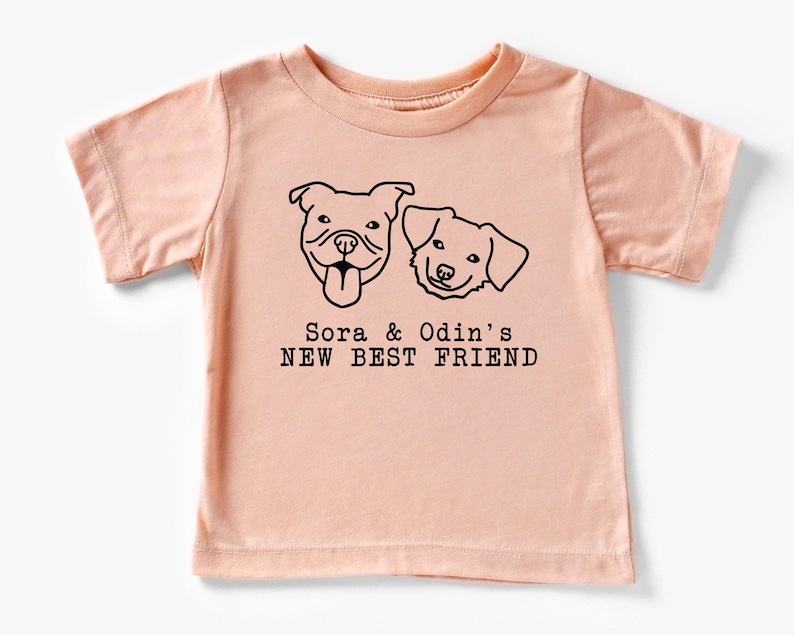 A light pink t-shirt with a graphic design featuring two dog faces and the text "Sora & Odin's NEW BEST FRIEND".