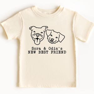A white t-shirt with a graphic design featuring two dog faces and the text "Sora & Odin's NEW BEST FRIEND".