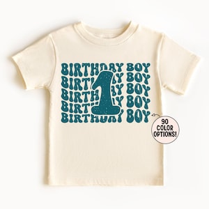 Może przedstawiać: Kremowy t-shirt z krótkim rękawem z powtarzającym się turkusowym nadrukiem graficznym z napisem "BIRTHDAY BOY" i dużą cyfrą "1" pośrodku. Mała okrągła odznaka w prawym dolnym rogu zawiera napis "90 COLOR OPTIONS!"