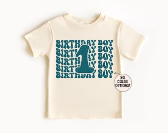 May include: A cream-colored short-sleeve t-shirt featuring a repeating teal graphic print of the text "BIRTHDAY BOY" with a large number "1" in the center. A small circular badge on the lower right displays the text "90 COLOR OPTIONS!"
