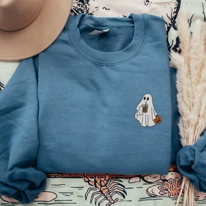 May include: A blue sweatshirt with a white ghost holding a cup of coffee and a small orange pumpkin. The sweatshirt is folded and laid on a patterned fabric.