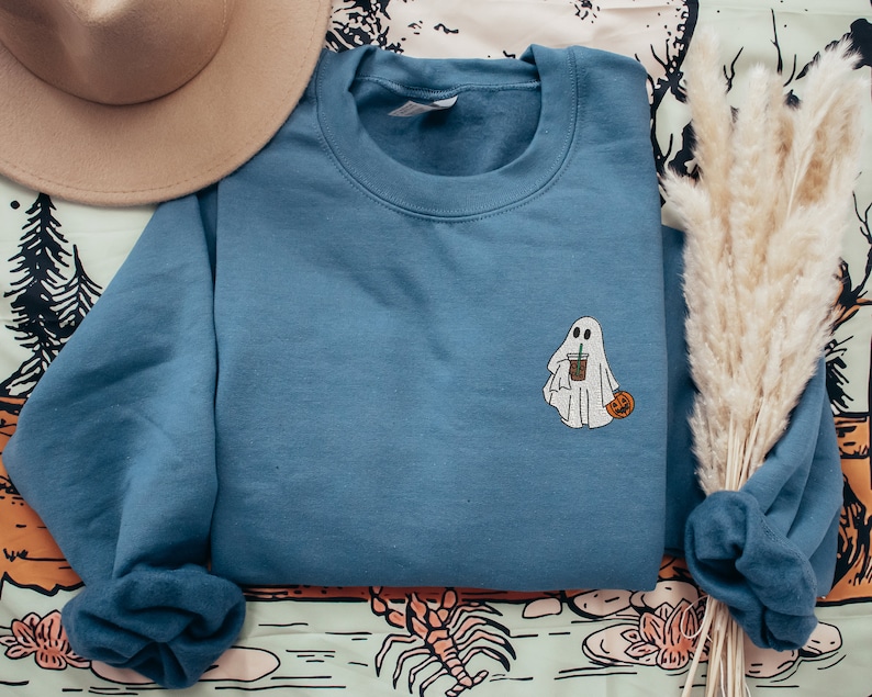 May include: A blue sweatshirt with a white ghost holding a cup of coffee and a small orange pumpkin. The sweatshirt is folded and laid on a patterned fabric.