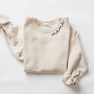 Op de afbeelding: Een cr&egrave;mekleurige sweatshirt met het woord "mama" in zwart script gedrukt op de linkerbovenborst. De lange mouwen zijn opgerold. De sweatshirt is gemaakt van een zacht, comfortabel materiaal.