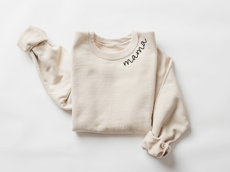 Op de afbeelding: Een cr&egrave;mekleurige sweatshirt met het woord "mama" in zwart script gedrukt op de linkerbovenborst. De lange mouwen zijn opgerold. De sweatshirt is gemaakt van een zacht, comfortabel materiaal.