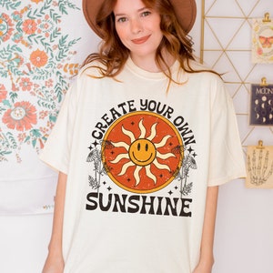 K&ouml;nnte beinhalten: Ein cremefarbenes T-Shirt mit einer Vintage-Grafik einer l&auml;chelnden Sonne mit dem Text "Create Your Own Sunshine" in Schwarz.