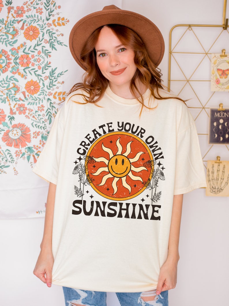 K&ouml;nnte beinhalten: Ein cremefarbenes T-Shirt mit einer Vintage-Grafik einer l&auml;chelnden Sonne mit dem Text "Create Your Own Sunshine" in Schwarz.