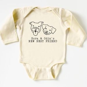 A baby onesie with a graphic of two dogs and the text "Sora & Odin's NEW BEST FRIEND" printed on it.