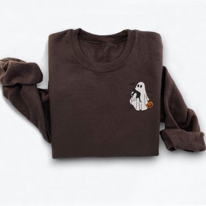 Op de afbeelding: Een bruin sweatshirt met een wit geborduurd spook dat een pompoen vasthoudt. Het spook wordt omringd door kleine zwarte vleermuizen.