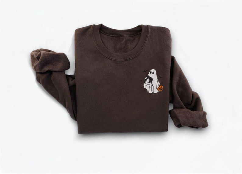 Op de afbeelding: Een bruin sweatshirt met een wit geborduurd spook dat een pompoen vasthoudt. Het spook wordt omringd door kleine zwarte vleermuizen.