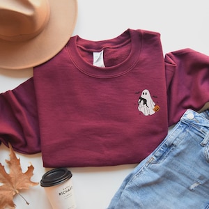Op de afbeelding: Een bordeauxrood sweatshirt met een geborduurd ontwerp van een geest die een zwarte kat en een pompoen vasthoudt. Het ontwerp bevindt zich op de voorkant van het sweatshirt.